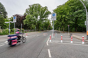 Kurierdienst mit einem Lastenrad auf einer Fahrradstraße mit seitlichen Grünmarkierungen, Piktogrammen und teilweise Pollern und Fahrradabstellbügeln.