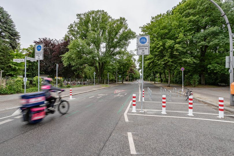 Kurierdienst mit einem Lastenrad auf einer Fahrradstraße mit seitlichen Grünmarkierungen, Piktogrammen und teilweise Pollern und Fahrradabstellbügeln.