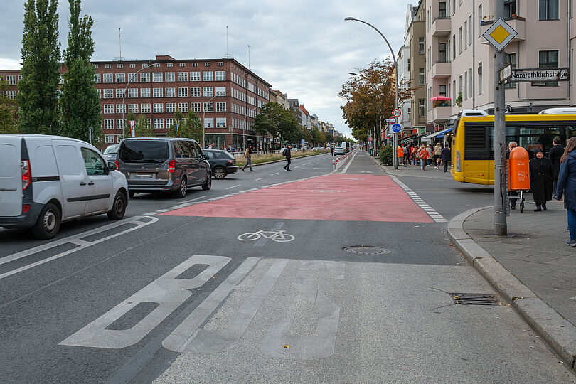 geschützter Radfahrstreifen am rechten Fahrbahnrand, daneben zwei Fahrspuren, auf einer belebten Geschäftsstraße, breite Rotmarkierung an einer Kreuzung zur Nebenstraße, Busspur