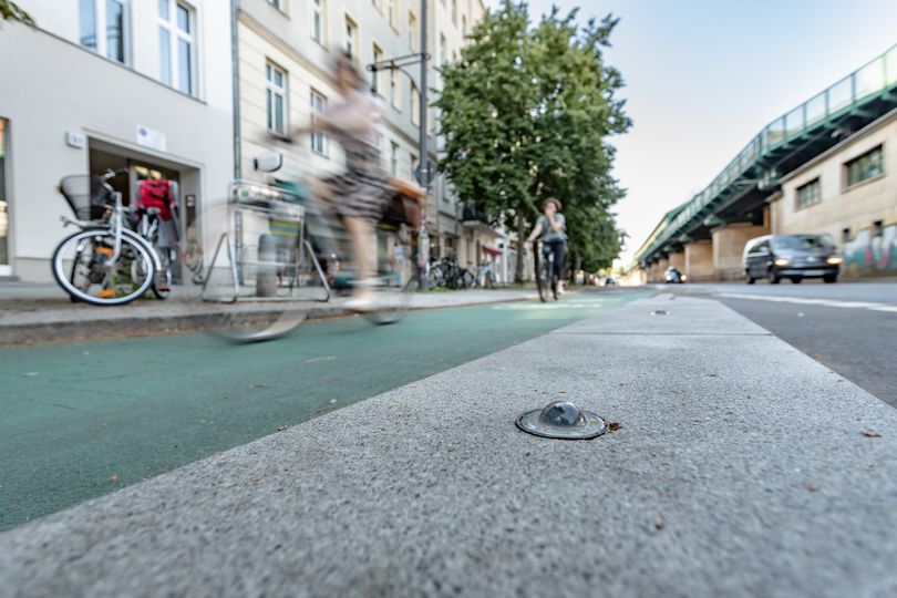 Nahaufnahme eines Betonbordes, die den Radfahrstreifen auf der Schönhauser Allee abtrennen. Ein Glasmarker auf dem Bord ist zu sehen. Auf dem grün markierten Radfahrstreifen fahren Fahrradfahrer.