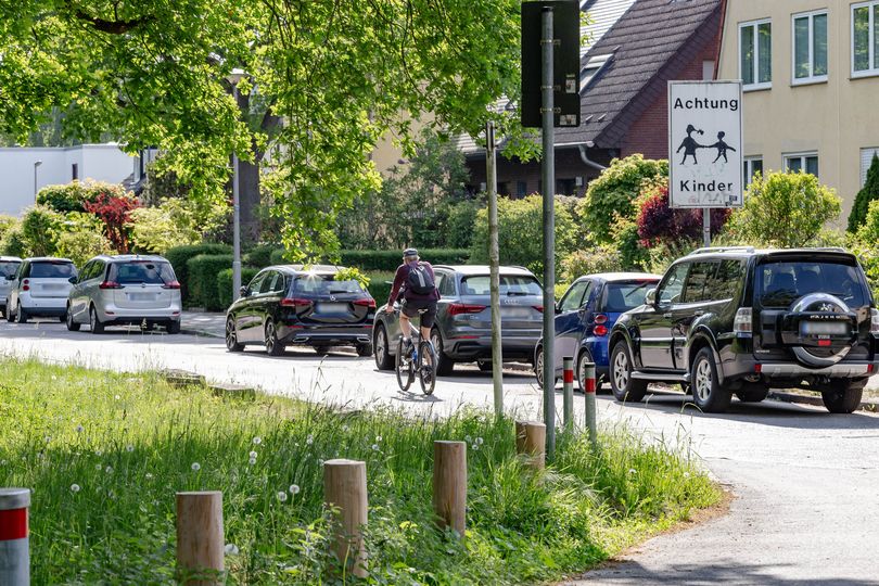 Wohnstraße mit parkenden Autos auf der rechten Fahrbahnseite, Radfahrer, Schild mit der Aufschrift "Achtung Kinder"