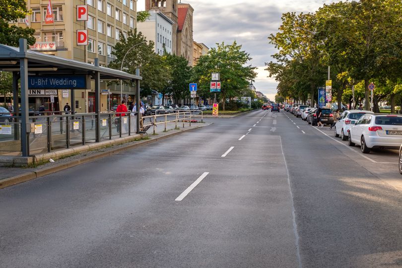zweispurige Straße in Mitte, rechts parkende Autos, links Mittelinsel mit U-Bahn-Eingang