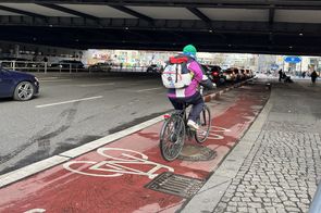 Radfahrende auf einem rotbeschichten Radfahrstreifen. Der Radfahrstreifen ist mit rotweiß markierten Trennelementen vom Autoverkehr getrennt.