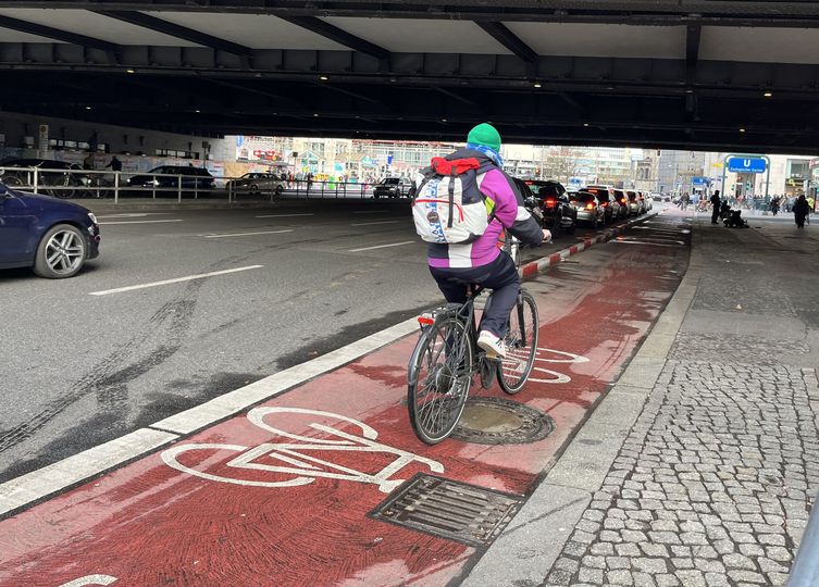 Radfahrende auf einem rotbeschichten Radfahrstreifen. Der Radfahrstreifen ist mit rotweiß markierten Trennelementen vom Autoverkehr getrennt.