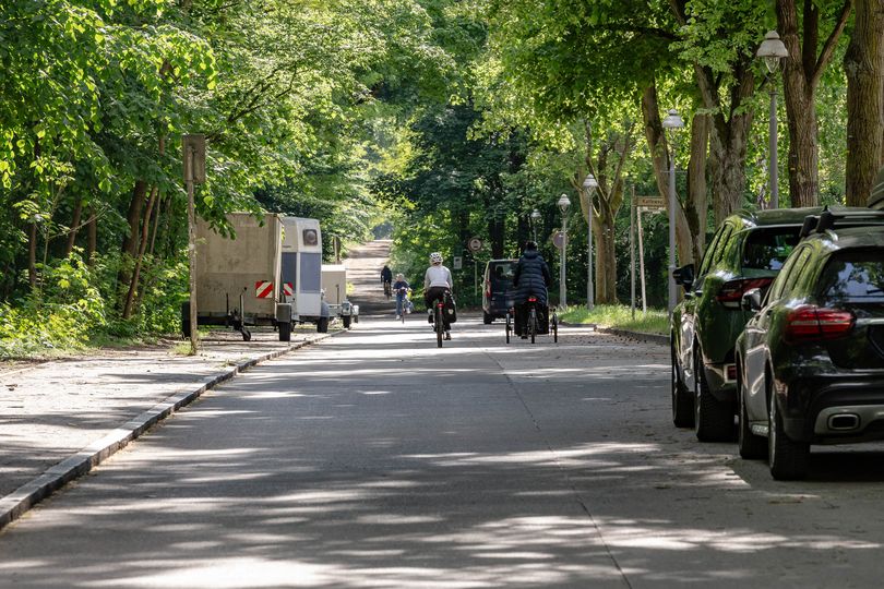 Radfahrende auf einer asphaltierten Straße mit parkenden Autos und Anhängern auf beiden Straßenseiten.