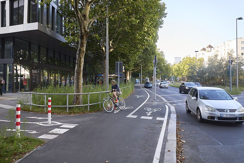 Hochboardradweg in einem Kreuzungsbereich mit unterschiedlichen Wegeführungen und Pollern mit Radfahrenden.