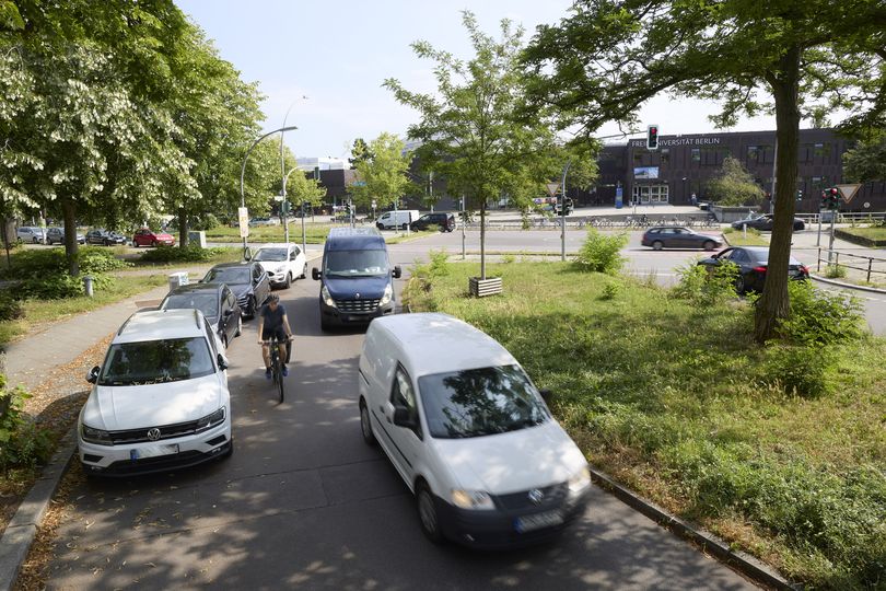 Straße in Berlin mit Autos, Radfahrer und Grünstreifen vor dem Gebäude der Freien Universität Berlin – urbaner Verkehr, eingeschränkte Verkehrssicherheit und mangelnde Radwege im Hochschulumfeld bei sonnigem Wetter.