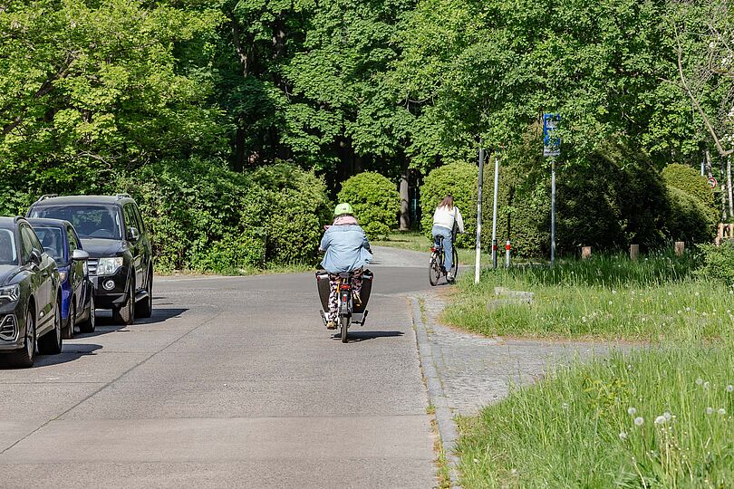 Radfahrende mit einem Lastenrad auf einer asphaltierten Straße in einem Wohngebiet.