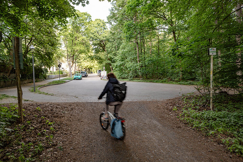 Radfahrende auf einem Waldweg, der zu einer asphaltierten Straße durch einen Wohngebiet führt.